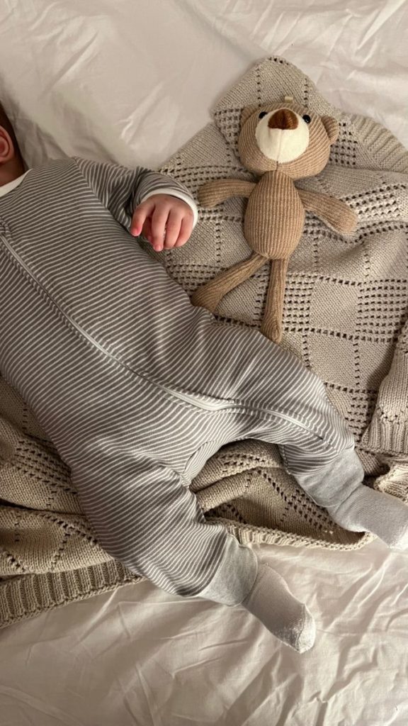 baby laying on bed in a neutral onsie, teddy bear and knit blanket.